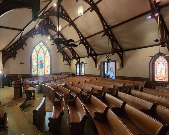 The image depicts the interior of a church with wooden pews, stained glass windows, and a vaulted ceiling supported by wooden beams.