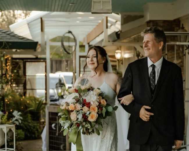 A bride in a white dress walks down a porch holding a bouquet of flowers, accompanied by a man in a black suit.