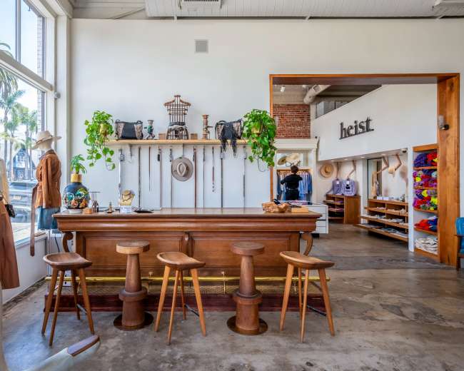 The interior of a clothing store features a wooden counter with bar stools, clothing displays on mannequins, and a variety of accessories arranged on shelves.