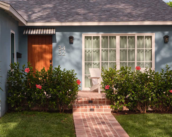 The image shows a blue house with a wooden door, white chairs on the porch, and a pathway lined with greenery and pink flowers.
