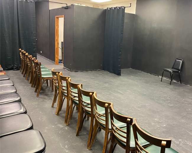 The image shows a sparsely furnished rehearsal room with two rows of wooden chairs facing a black wall, and two black curtains dividing the space.