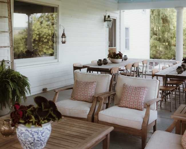 A covered porch features wooden furniture, potted plants, and large windows overlooking a green landscape.