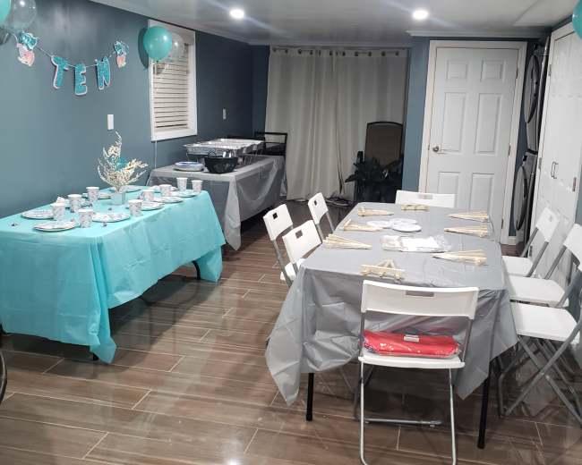 The image shows a party setup in a basement with tables covered in light blue and gray tablecloths, decorated with balloons and banners.