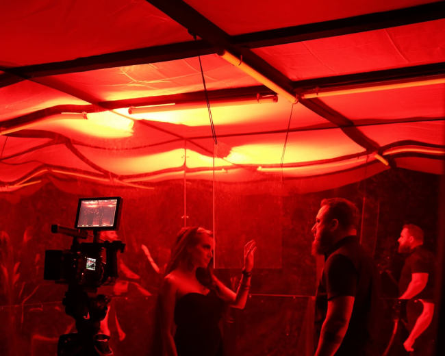 A woman in a black dress gestures toward a man in a black shirt under red lighting in a partially draped room.