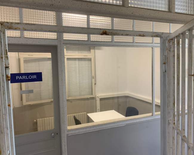 The image shows a view through a metal gate into a small, empty room with a table and a chair, labeled "PARLOIR."