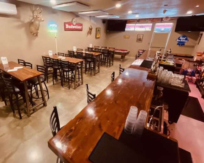 The image shows a spacious bar area with wooden tables and chairs, a long wooden bar counter, and various signs on the walls, including beer advertisements.