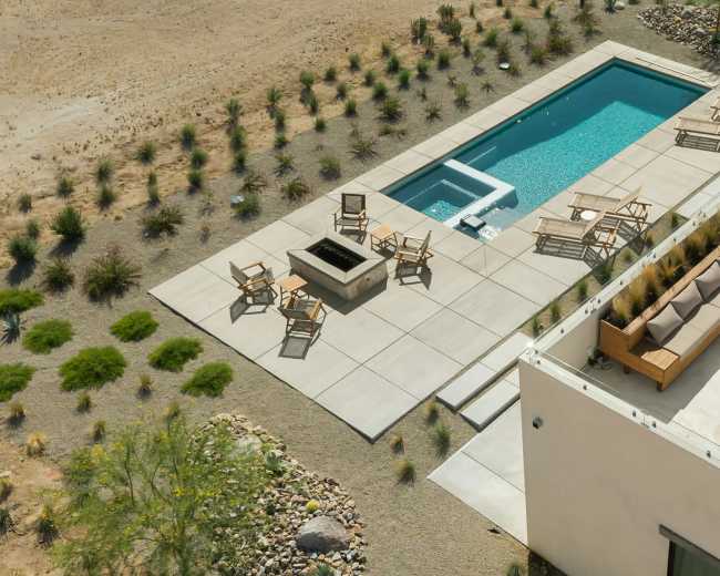 The image shows a modern patio area with a swimming pool, seating arrangements, and a fire pit, set in a desert landscape.