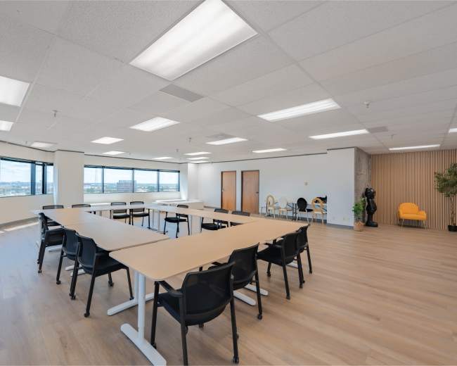 A large, bright meeting room features a long table surrounded by black chairs, with windows offering a view of the outside and minimalist decor on the walls.