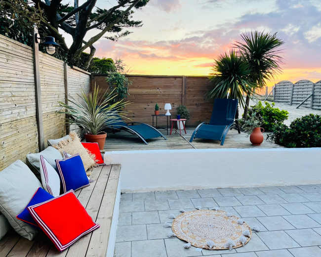 The image shows a cozy outdoor seating area with lounge chairs, potted plants, and decorative pillows, set against a colorful sunset sky.