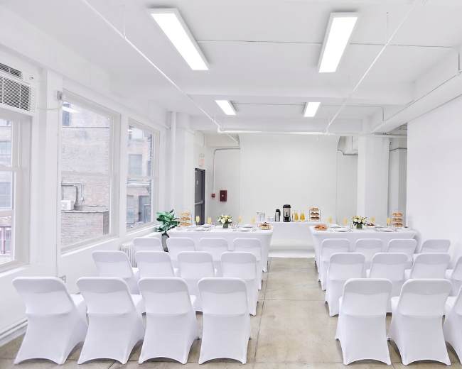 A bright, modern room features rows of white chairs facing a table set with refreshments, including coffee and pastries.
