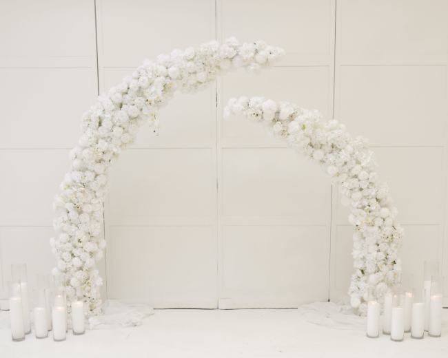 The image shows a floral arch made of white flowers arranged in two curved structures, with white candles placed on the ground nearby.