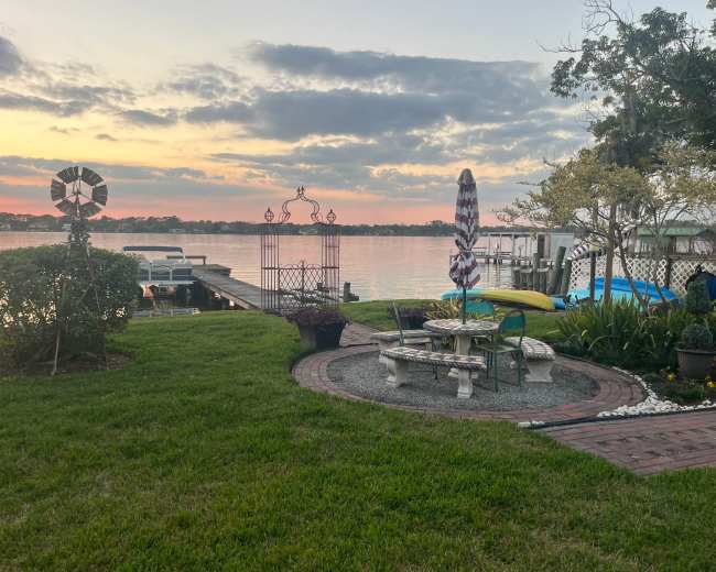 The scene features a waterfront with boats docked, a grassy area with a seating arrangement surrounded by decorative features, and a sunset sky in the background.