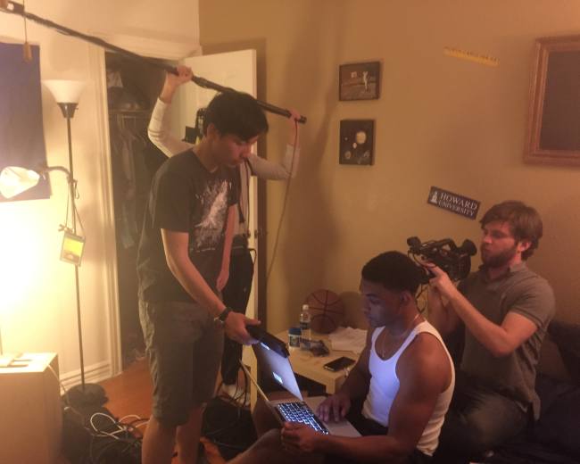 A film crew sets up equipment in a small room as one person operates a laptop while others adjust the lighting and camera.