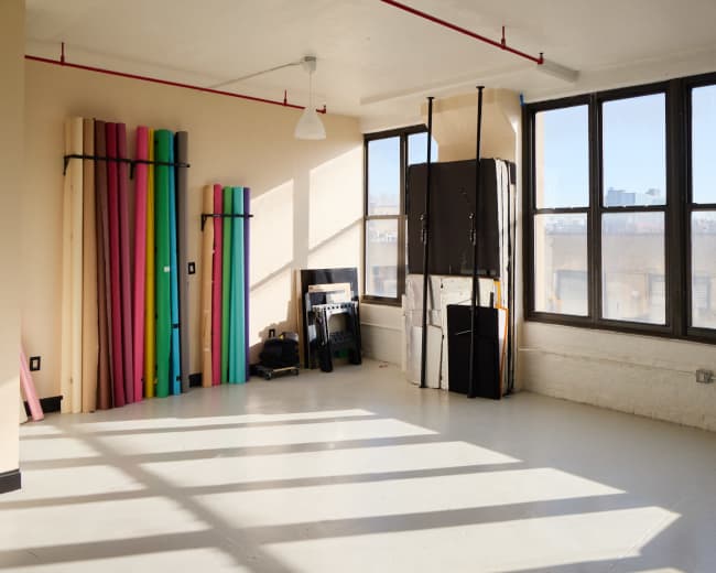The image shows an empty room with large windows, colorful rolls of paper stacked against one wall, and various equipment leaning against another.