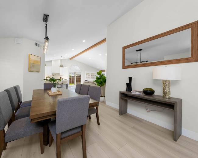 The image shows a modern dining area with a wooden table and gray upholstered chairs, set against a light-colored wall and a window that lets in natural light.