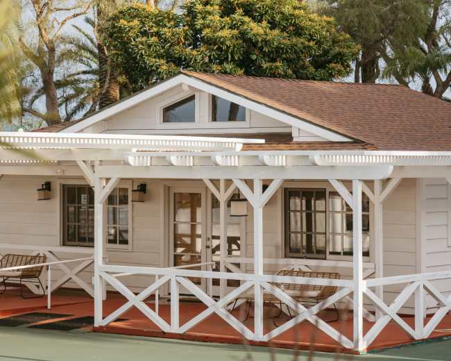 A small white cottage with a brown roof features a wraparound porch surrounded by greenery.