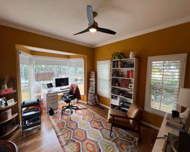 A home office with a desk, a black office chair, a brown armchair, shelves filled with books, and large windows providing natural light.