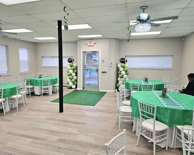 The image shows a decorated event space featuring green tablecloths, white chairs, and an entrance adorned with balloon columns.
