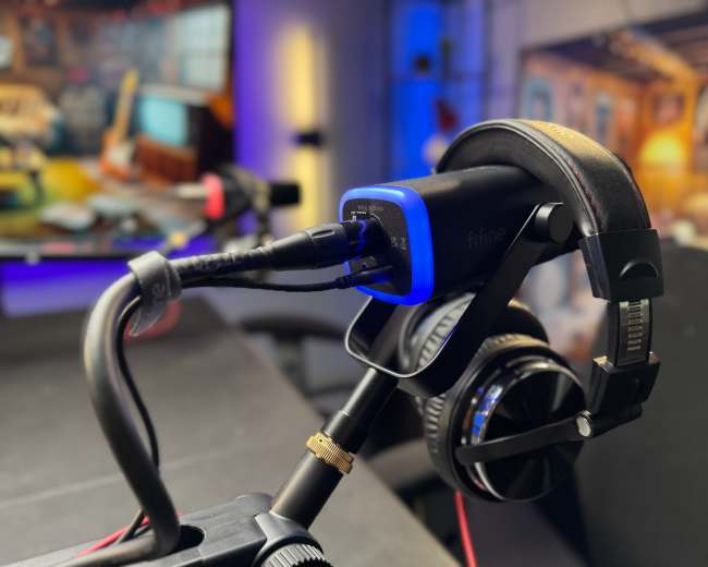 A microphone and headphones are positioned on a stand in front of a blurred background featuring lighting and equipment typically found in a studio setting.