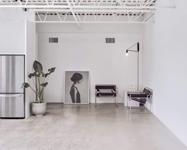 A minimalist interior features a large black-and-white portrait on the floor, a potted plant, a metal chair, and a metal side chair, all set against a white wall.