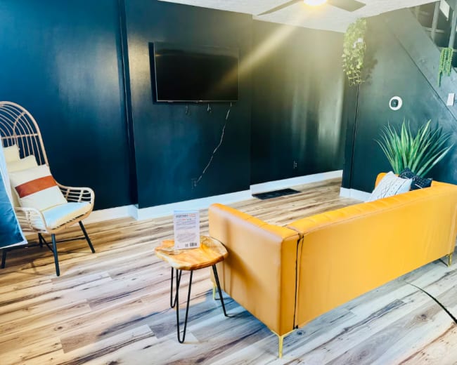 The image shows a living room with a yellow couch, a small wooden side table, a modern chair, a television on the wall, and minimal greenery.
