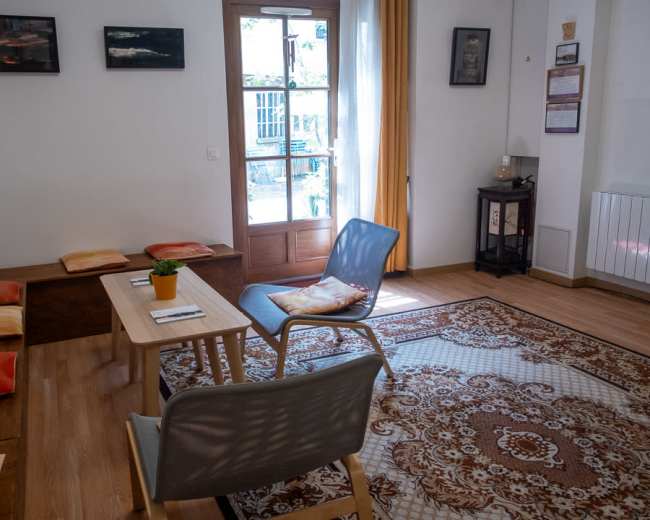 The image shows a cozy room with a wooden table and chairs, a patterned area rug, and a large window that lets in natural light.