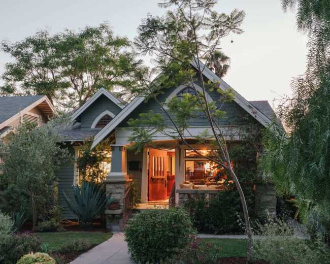 A craftsman-style house with a welcoming front porch is illuminated at dusk, surrounded by lush greenery and landscaping.