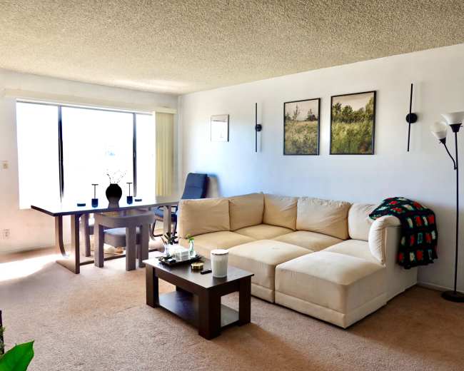 The image shows a living room with a beige sectional sofa, a small wooden coffee table, a dining table with chairs, and framed artwork on the wall.