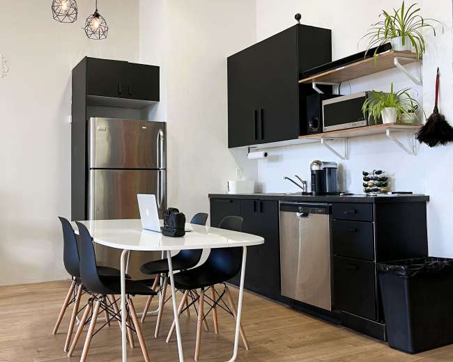 The image shows a modern kitchen with black cabinetry, stainless steel appliances, a white table with black chairs, and pendant lighting.