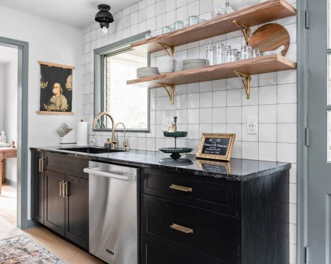 A modern kitchen featuring black cabinetry, a stainless steel sink, and open wooden shelving with glassware against a tiled wall.