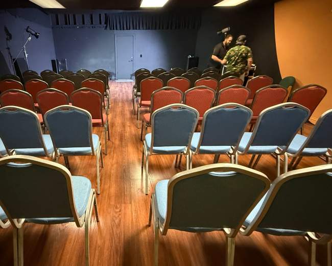 The image shows rows of empty chairs in a performance space with two people preparing in the background.