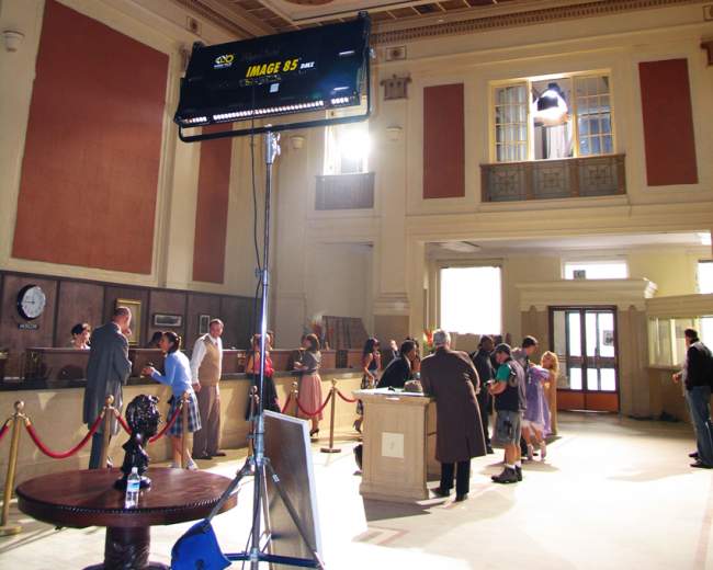 A film crew sets up a scene inside a grand, partially lit hall featuring high ceilings and decorative architecture.