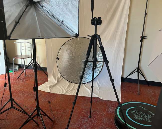 The image shows a photography setup featuring softbox lights, a camera on a tripod, a reflector, and a white backdrop in a studio environment.