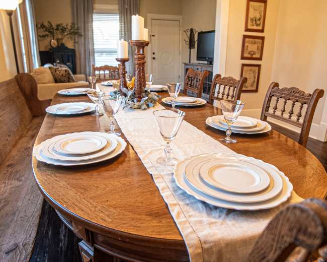 A large wooden dining table is set with white plates and glassware, surrounded by wooden chairs, in a well-decorated dining room.