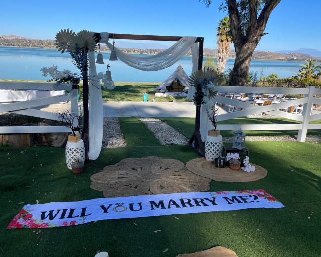 The scene features a wedding proposal setup by a lake, adorned with a decorative arch, a large floral banner reading "WILL YOU MARRY ME?", and carefully arranged decorations around a green lawn.