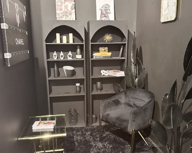 The image shows a small, dark room with a black wall, featuring a corner bookshelf, a velvet chair, a glass side table, and decorative items.