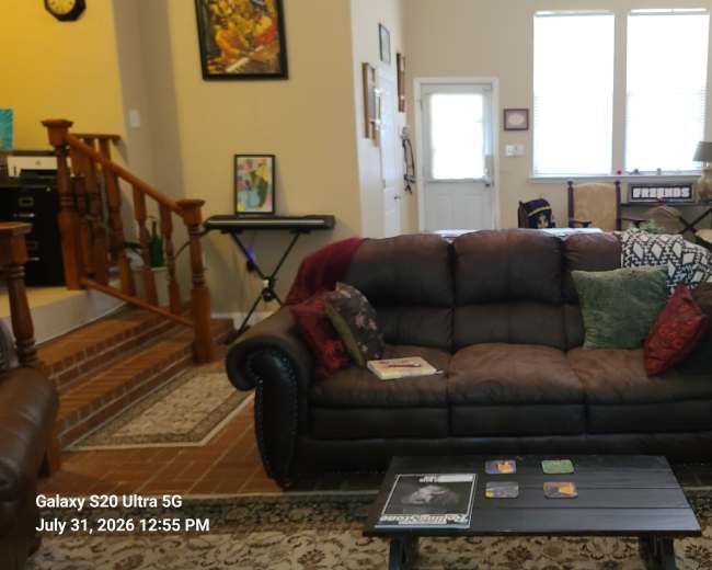 The image shows a cozy living room with a brown leather couch, a coffee table, and various decorative items and furniture arranged around the space.