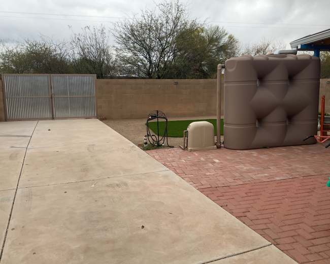 The image shows a fenced outdoor area with a concrete and brick path, featuring a large tan water tank and a patch of artificial grass.