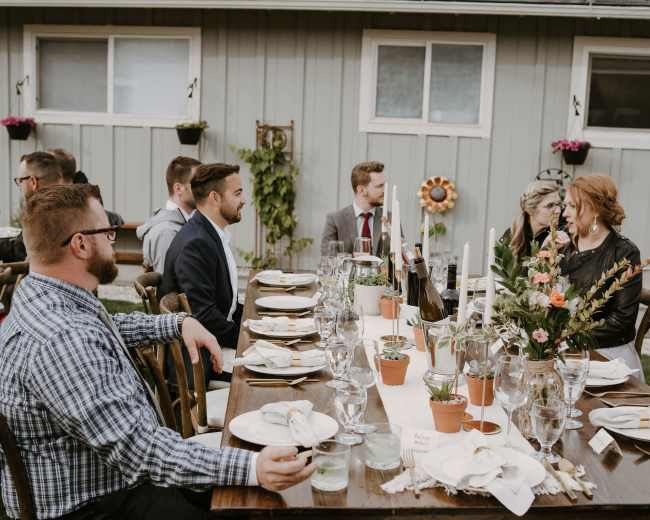 A group of elegantly dressed people sits around a long wooden table set for dinner in a garden setting.