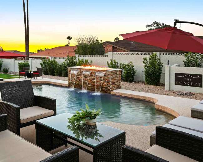 The image shows a modern outdoor lounging area beside a pool with a stone fountain and red umbrellas, set against a colorful sunset.