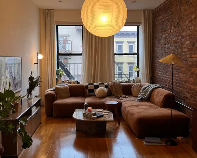 A cozy living room features a brown sectional sofa, a round coffee table, and large windows framed by light curtains.