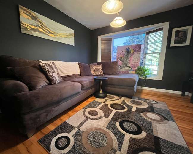The image shows a cozy living room with a large sectional sofa, a circular coffee table, and a patterned area rug, complemented by a window and wall art.