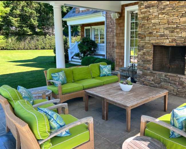 A patio area features green cushioned wicker furniture arranged around a wooden coffee table in front of a stone fireplace.