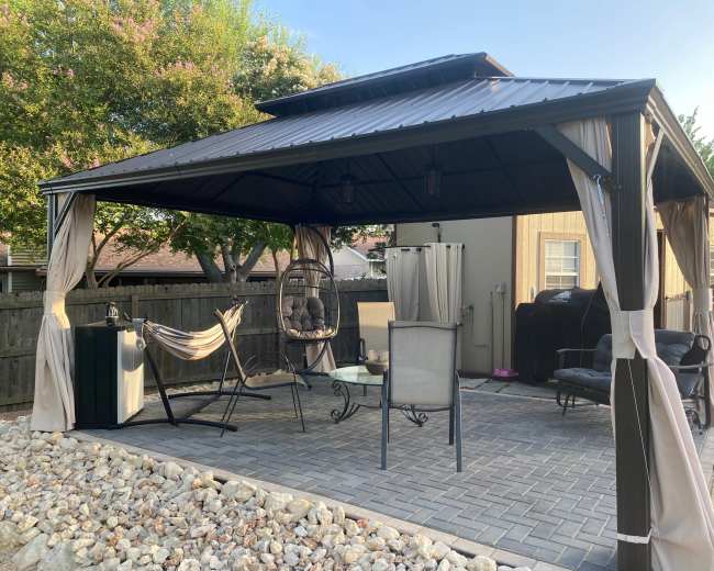 A metal gazebo with a high roof is furnished with several chairs and a hanging swing, surrounded by a stone patio and trees in the background.