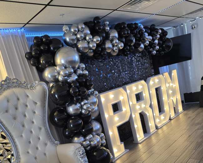 The scene features a decorative wall with a large "PROM" sign in lights, surrounded by a balloon arch in black and silver, with a white ornate chair nearby.