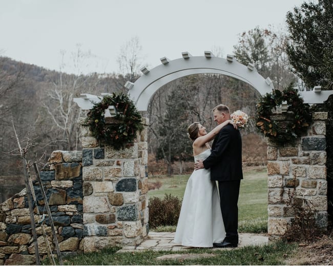 A couple embraces under a stone archway adorned with wreaths, set against a backdrop of trees and a lake.