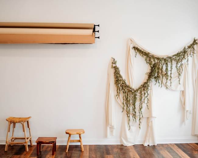 The image shows a minimalist interior featuring a white wall adorned with draped fabric and greenery, along with wooden stools positioned on the floor.