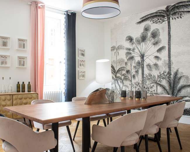 The image shows a modern dining room with a large wooden table surrounded by upholstered chairs, a light fixture overhead, and a decorative mural featuring palm trees on one wall.