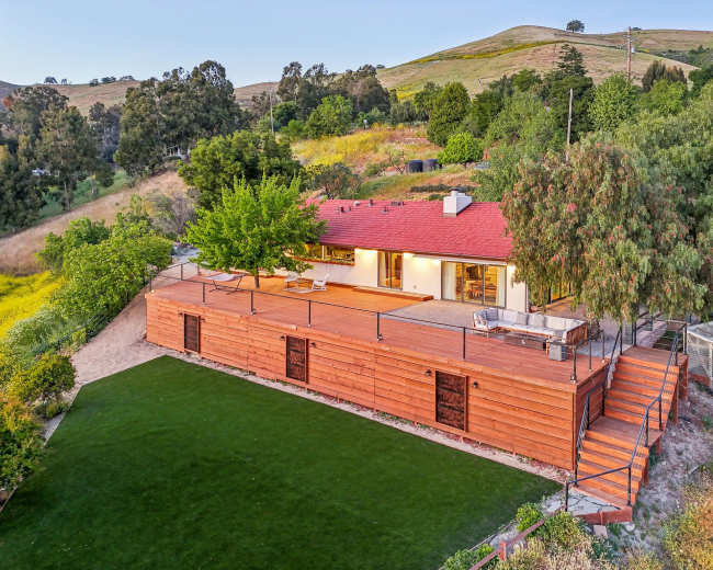 A modern single-story house with a red roof and wooden exterior sits on a hillside surrounded by greenery and a landscaped lawn.