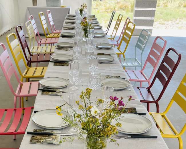 A long dining table is set with plates and glassware, surrounded by colorful chairs and decorated with a small vase of flowers.